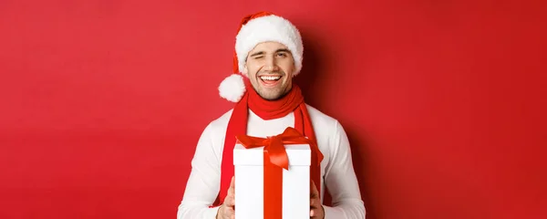 Pojęcie ferii zimowych, Bożego Narodzenia i stylu życia. Przystojny bezczelny mężczyzna w czapce i szaliku Mikołaja, trzymający obecny i uśmiechnięty, mrugający przed kamerą, stojący nad czerwonym tłem — Zdjęcie stockowe