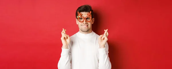 Närbild av stilig nervös kille i festglasögon, gör en önskan, korsa fingrar för lycka till och tittar med hopp på kameran, står över röd bakgrund — Stockfoto
