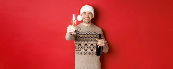 Begrip wintervakantie, nieuwjaar en viering. Portret van een knappe man met kerstmuts en trui, champagne vasthoudend, glas hijgend en proost zeggend op kerstfeest — Stockfoto