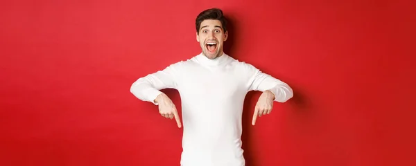 Изображение изумленного красивого парня в белом свитере, реагирующего на рождественскую рекламу, указывающего пальцами вниз, показывающего логотип, стоящего на красном фоне — стоковое фото