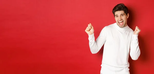 Portrait of handsome man enjoying new year party, dancing and having fun, standing in white sweater against red background — Stock Photo, Image