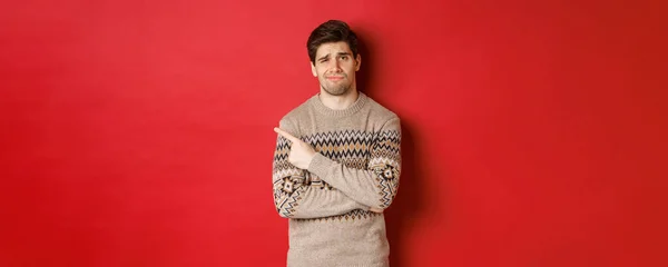 Imagem do homem cético e descontente em camisola de Natal, apontando para o canto superior esquerdo sem diversão, mostrando mau presente de Natal, de pé sobre fundo vermelho — Fotografia de Stock