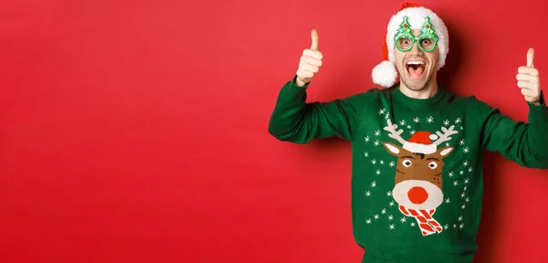Retrato de jovem super feliz em óculos de festa, chapéu de santa e suéter, mostrando polegar para cima na aprovação, recomendando oferta promocional ano novo, de pé sobre fundo vermelho — Fotografia de Stock