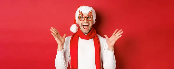 Concept of christmas, winter holidays and celebration. Image of surprised and happy man looking amazed, wearing party glasses and enjoying new year, standing over red background — Stock Photo, Image