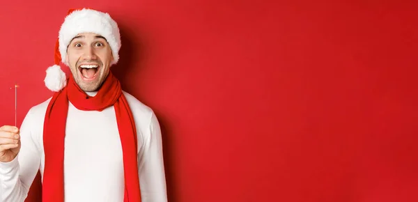 Концепция Рождества, зимних праздников и празднования. Крупный план счастливого красавца в шляпе Санты и шарфе, веселящегося на новогодней вечеринке, держащего искру и улыбающегося, красный фон — стоковое фото