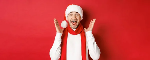 Концепция Рождества, зимних праздников и празднования. Изображение красивого мужчины, удивлённого новогодним промо-предложением, улыбающегося удивлённого, стоящего на красном фоне — стоковое фото