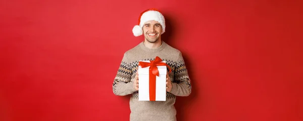 Изображение красивого улыбающегося мужчины в шляпе Санты и зимнем свитере, держащего подарок, дарящего рождественский подарок и желающего счастливых праздников, стоящего на красном фоне — стоковое фото