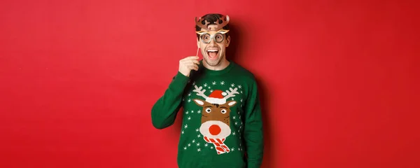 Hombre guapo sorprendido en suéter de Navidad, celebración de la máscara de fiesta y sonriendo, disfrutando de la celebración del año nuevo, de pie sobre el fondo rojo — Foto de Stock