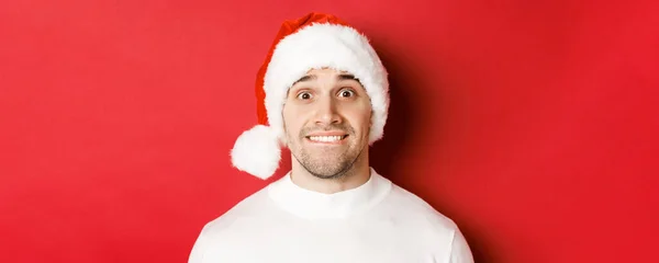 Close-up van knappe man in kerstmuts, bijtende lip en kijkend met verleiding naar iets wat hij wil, staande over rode achtergrond — Stockfoto