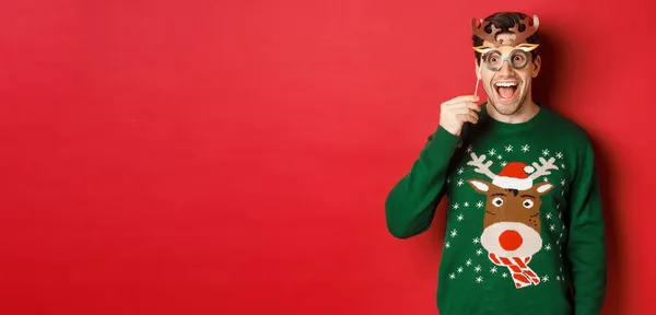 Bonito homem surpreso em camisola de Natal, segurando máscara de festa e sorrindo, desfrutando de celebração do ano novo, de pé sobre fundo vermelho — Fotografia de Stock