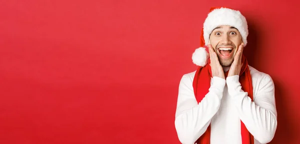 Концепция Рождества, зимних праздников и празднования. Крупный план удивленного и счастливого человека в шляпе Санты и шарфе, смотрящего на что-то удивительное, стоящего на красном фоне — стоковое фото