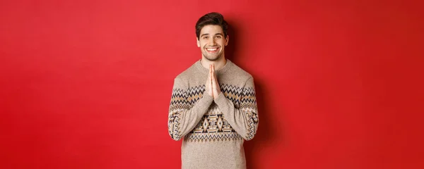 Образ щасливого і милого чоловіка в різдвяному светрі, просячи про послугу, тримаючи руки разом, посміхаючись і кажучи спасибі, відчуваючи себе вдячним, стоячи на червоному тлі — стокове фото