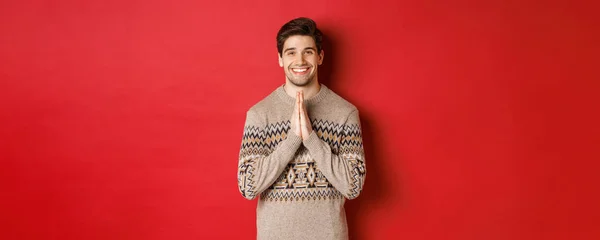 Imagen de hombre feliz y lindo en jersey de navidad, pidiendo favor, tomados de la mano, sonriendo y diciendo gracias, sintiéndose agradecido, de pie sobre fondo rojo —  Fotos de Stock