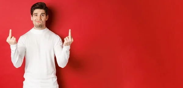 Зображення засмученого і засмученого чоловіка, який каже трахатись, показуючи середні пальці і дивлячись засмучений, стоїть на червоному тлі в білому светрі — стокове фото