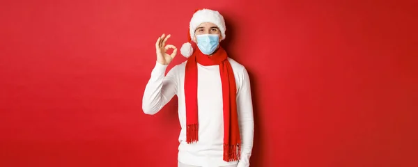 Covid-19, noel ve salgın sırasında tatil kavramı. Tıbbi maske ve Noel Baba şapkası takan mutlu ve memnun bir adamın portresi onay işareti gösteriyor, bir şey öneriyor. — Stok fotoğraf