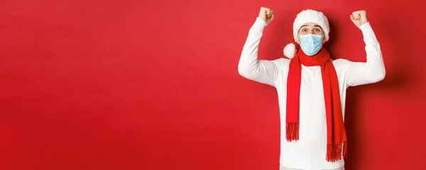 Concept covid-19, karácsony és ünnepek világjárvány idején. Portré boldog ember télapó kalap és orvosi maszk, öröm és ünnepli az új évet, áll a piros háttér felett — Stock Fotó