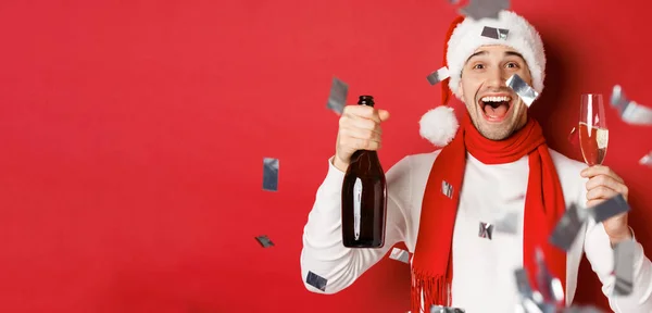 Концепция зимних праздников, Рождества и образа жизни. Крупный план счастливого человека, празднующего Новый год, держащего бутылку шампанского и бокал, стоящего на красном фоне с конфетти — стоковое фото