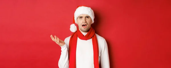 クリスマス、冬の休日やお祝いの概念。サンタの帽子とスカーフで混乱した男の肖像、眉をひそめ、複雑に見える、赤い背景の上に立って — ストック写真