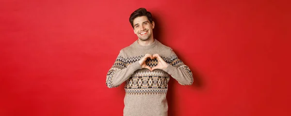 Kuva komea parrakas kaveri talvella villapaita, toivottaa hyvää joulua ja osoittaa sydämen merkki, hymyillen ihana kameralle, seisoo punaista taustaa vasten — kuvapankkivalokuva
