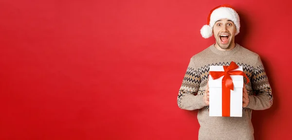 Obraz podekscytowanego przystojnego mężczyzny otrzymującego prezent świąteczny, noszącego czapkę Mikołaja i sweter zimowy, krzyczący z radości, trzymający prezent i stojący nad czerwonym tłem — Zdjęcie stockowe