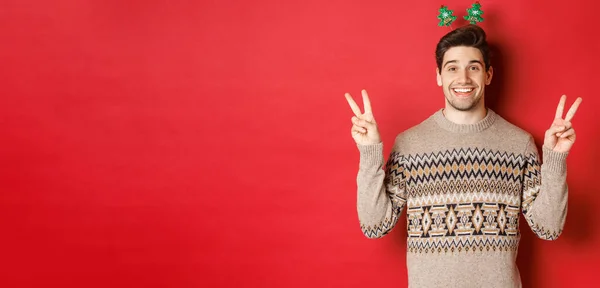 Концепция зимних праздников, Рождества и праздника. Изображение красивого и глупого парня, одетого для новогодней вечеринки, показывающего знаки мира и улыбающегося, стоящего на красном фоне — стоковое фото