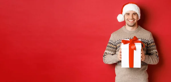 Bild eines gut aussehenden lächelnden Mannes mit Weihnachtsmütze und Winterpullover, der ein Geschenk in der Hand hält, Weihnachtsgeschenk macht und frohe Feiertage wünscht, vor rotem Hintergrund stehend — Stockfoto
