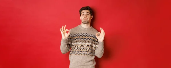 stock image Impressed handsome man in christmas sweater, showing okay signs and praising good party, nodding in approval, standing pleased against red background