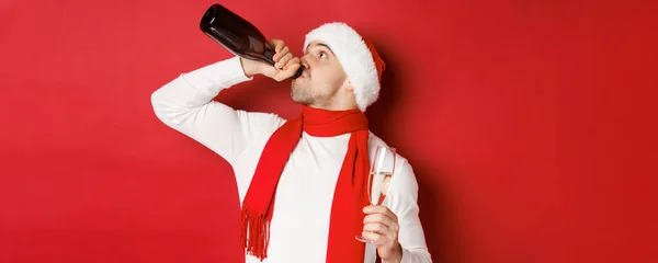 Kış tatili, Noel ve yaşam tarzı kavramı. Yeni yıl partisinde sarhoş olan, şişeden şampanya içen, Noel Baba şapkası takan, kırmızı arka planda duran bir adam. — Stok fotoğraf