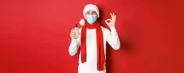 Covid-19, noel ve salgın sırasında tatil kavramı. Noel Baba şapkası ve tıbbi maskeli çekici bir adam. El dezenfektanı tavsiye ederken, kırmızı arka planda duruyor. — Stok fotoğraf