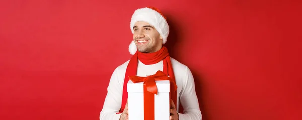 Concept de vacances d'hiver, Noël et style de vie. Gros plan de gars attrayant dans le chapeau et l'écharpe du Père Noël, souriant et regardant à gauche tout en tenant le nouvel an présent, debout sur fond rouge — Photo