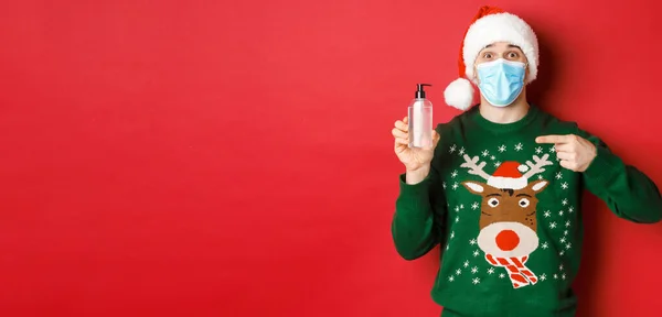 Концепція нового року, коронавірус та соціальне дистанціювання. Портрет веселого чоловіка в капелюсі Санта, різдвяний светр і медична маска, вказуючи пальцем під рукою дезінфікуючий засіб, стоїть на червоному тлі — стокове фото