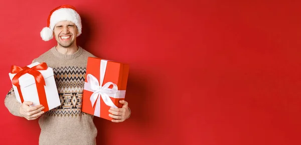 Koncepcja ferii zimowych, Nowy Rok i świętowanie. Obraz szczęśliwy i podekscytowany młody człowiek lubi prezenty, trzymając prezenty i uśmiechnięty, noszenie Santa kapelusz i sweter świąteczny — Zdjęcie stockowe