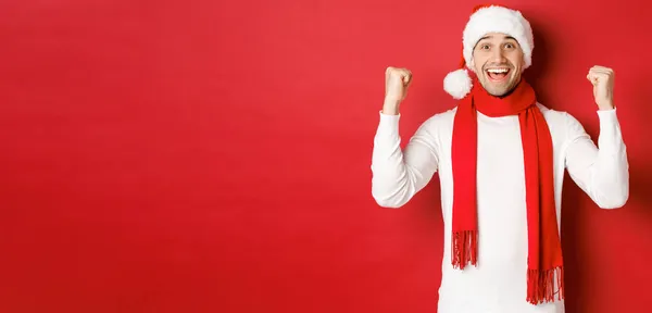 Retrato de homem feliz e animado em santa chapéu e cachecol, alegrando-se e ganhar algo, celebrando o ano novo, de pé sobre fundo vermelho — Fotografia de Stock