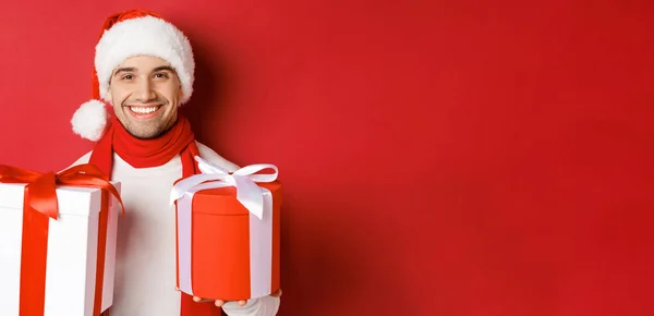 Begrip wintervakantie, kerst en levensstijl. Close-up van glimlachende knappe man in santa hoed en sjaal, wensen gelukkig nieuwjaar en het houden van geschenken, staan met cadeautjes over rode achtergrond — Stockfoto