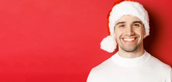 Primer plano del atractivo hombre sonriente en jersey blanco y sombrero de santa, luciendo feliz, disfrutando de las vacaciones de invierno, de pie sobre fondo rojo —  Fotos de Stock