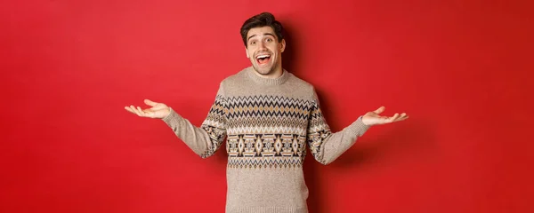 Портрет щасливого і здивованого, красивого кавказького хлопця, одягненого в різдвяний светр, розсунутий руками вбік і виглядає безглуздо, стоїть над червоним тлом — стокове фото