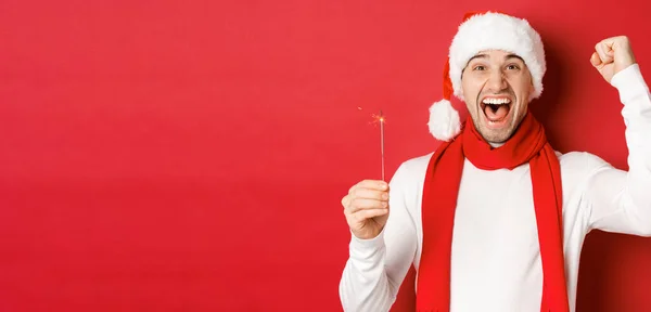 Noel, kış tatili ve kutlama kavramı. Heyecanlı yakışıklı adamın portresi, elini kaldırıp maytap tutuyor, mutlu yıllar diliyor, kırmızı arka planda duruyor. — Stok fotoğraf