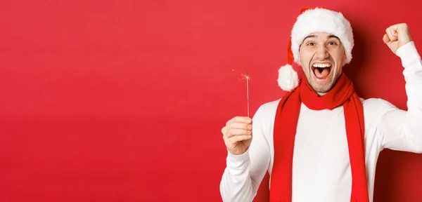 Concept of christmas, winter holidays and celebration. Portrait of excited handsome man, raising hand up and holding sparkler, wishing happy new year, standing over red background — Stock Photo, Image