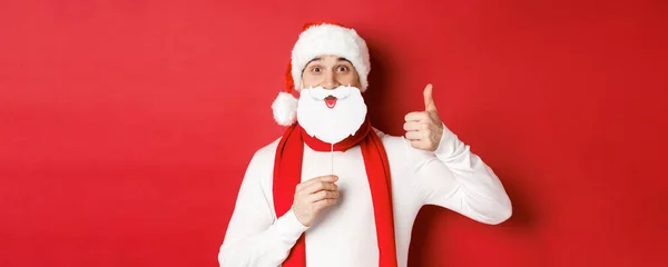 Έννοια των Χριστουγέννων, χειμερινές διακοπές και γιορτή. Πορτρέτο του αστείου άνδρα με το καπέλο santa, κρατώντας μάσκα γενειάδας, δείχνοντας τον αντίχειρα προς τα πάνω, απολαμβάνοντας το κόμμα της Πρωτοχρονιάς, στέκεται πάνω από το κόκκινο φόντο — Φωτογραφία Αρχείου