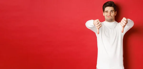 Portrait of disappointed skeptical guy, wearing white sweater, showing thumbs-down to disagree, express dislike, standing over red background — Stock Photo, Image