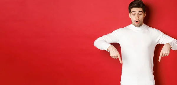 Portrait of surprised good-looking guy in white sweater, looking and pointing fingers down at logo, standing against red background — Stock Photo, Image