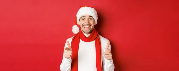 Koncepcja Bożego Narodzenia, ferie zimowe i uroczystości. Bezczelny mężczyzna w czapce i szaliku Mikołaja, uśmiechnięty i wytykający palcami przed kamerą, życzący szczęśliwego Nowego Roku, stojący nad czerwonym tłem — Zdjęcie stockowe