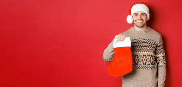 Konzept der Winterferien, Neujahr und Feier. Bild eines gut aussehenden lächelnden Mannes mit Weihnachtsmütze und Pullover, der Weihnachtsstrumpf für Geschenke und Bonbons in der Hand hält, vor rotem Hintergrund stehend — Stockfoto