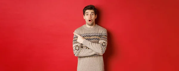 Afbeelding van onder de indruk knappe man, tonen ontzagwekkende kerst promo aanbod, wijzende vinger op de linker bovenhoek en kijken verbaasd, staan in xmas trui over rode achtergrond — Stockfoto
