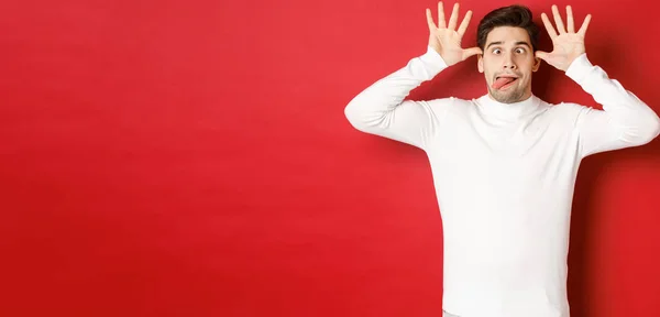 Retrato de cara caucasiano engraçado, mostrando língua e fazendo rostos, vestindo suéter branco, de pé contra fundo vermelho — Fotografia de Stock