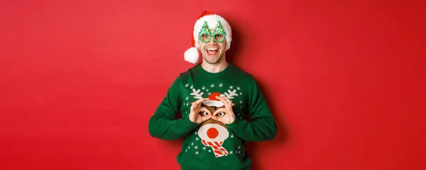 Πορτρέτο του ξέγνοιαστου όμορφου άντρα με το καπέλο και τα γυαλιά του, κοροϊδεύοντας το χριστουγεννιάτικο πουλόβερ του, δείχνοντας χαρούμενος πάνω από το κόκκινο φόντο — Φωτογραφία Αρχείου