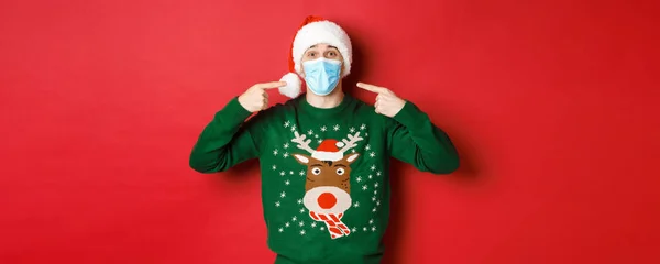 새해의 컨셉, 코로나 바이러스 그리고 사회적 거리. 산타 모자와 크리스마스 스웨터를 입고 빨간 배경 위에 서서 파티에서 의료용 마스크를 쓰기를 추천하는 행복 한 남자 — 스톡 사진