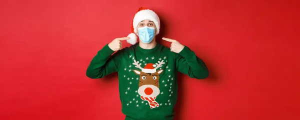 Conceito de ano novo, coronavírus e distanciamento social. Homem feliz em chapéu de Papai Noel e camisola de Natal, recomendando usar máscara médica na festa, de pé sobre fundo vermelho — Fotografia de Stock