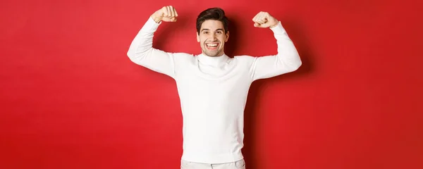 Portrét usmívajícího se pohledného muže v bílém svetru, ohýbání bicepsy a vychloubání silou, předvádění silné svaly po cvičení, stojící nad červeným pozadím — Stock fotografie
