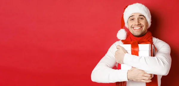 Begreppet vinter semester, jul och livsstil. Bild av härlig kille i Santa hatt och halsduk, krama hans nya år närvarande och leende smickrad, står över röd bakgrund — Stockfoto